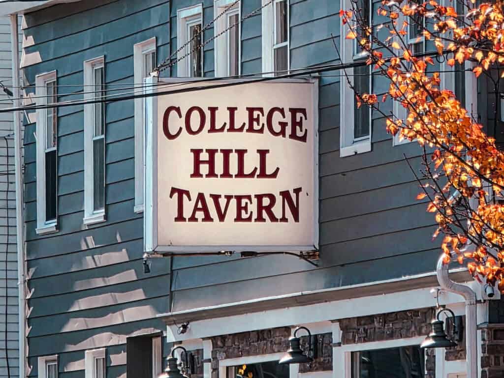 College Hill Tavern sign mounted on a blue building with windows above, orange tree leaves visible to the right, power lines crossing foreground, daytime lighting.