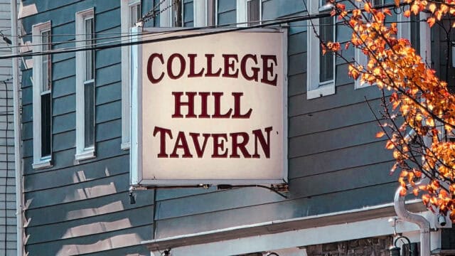 College Hill Tavern sign mounted on a blue building with windows above, orange tree leaves visible to the right, power lines crossing foreground, daytime lighting.