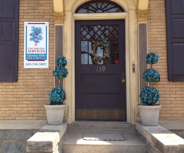 Front entrance of a building decorated with blue floral arrangements in large white pots, featuring a black door with a wreath and a sign for counseling services on the brick wall.