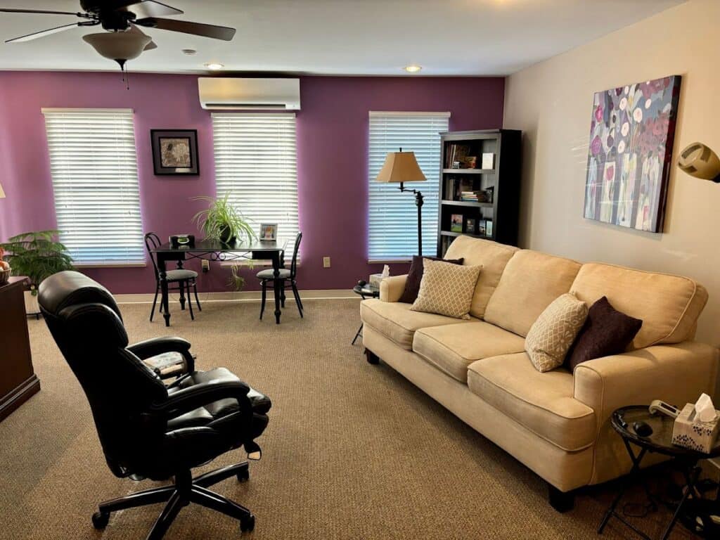Comfortable living room featuring a beige sofa, black office chair, purple accent wall, and natural light from windows, creating a welcoming and stylish space.