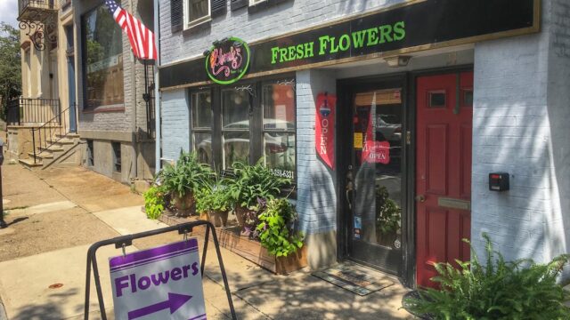 Exterior view of Bloomie's Flower Shop with fresh flowers outside, a purple flower sign, and a black and green 'Fresh Flowers' sign above the entrance.