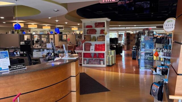 Interior view of a retail store with a curved wooden checkout counter, display shelves, and colorful luggage in a well-lit, modern shopping environment.