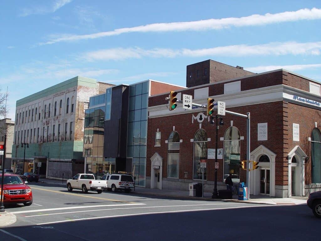 City street view showing a mix of historic and modern buildings, traffic lights, and cars parked along the curb under a blue sky with wispy clouds.