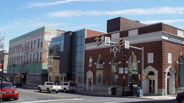 City street view showing a mix of historic and modern buildings, traffic lights, and cars parked along the curb under a blue sky with wispy clouds.