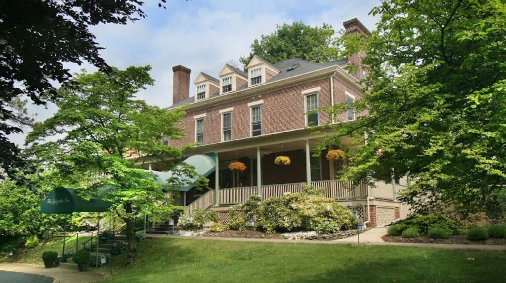 Historic brick Lafayette Inn surrounded by lush green trees and landscaped garden, featuring a spacious porch with hanging flower baskets and outdoor seating.