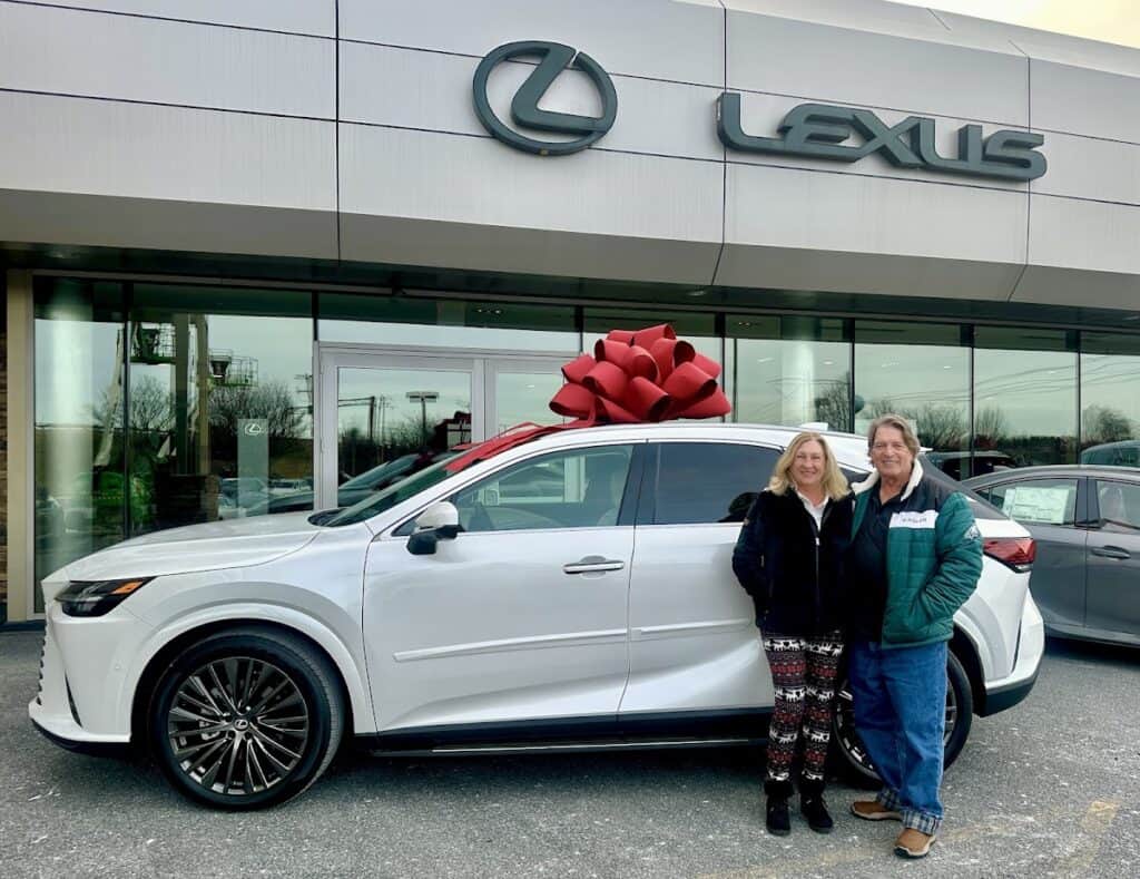 Happy couple standing next to a new white Lexus SUV with a large red bow in front of Lexus of Lehigh Valley dealership.