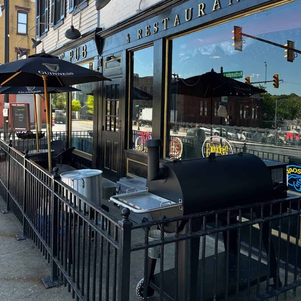 Outdoor seating area with a black grill and umbrellas outside Porters' Pub & Restaurant in Easton, featuring a sidewalk fence and street view.