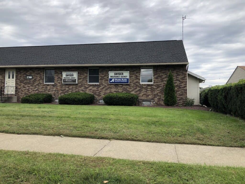 Snyder Insurance Agency building with brick exterior, three windows, and a sign, surrounded by a well-maintained lawn and bushes under a cloudy sky.