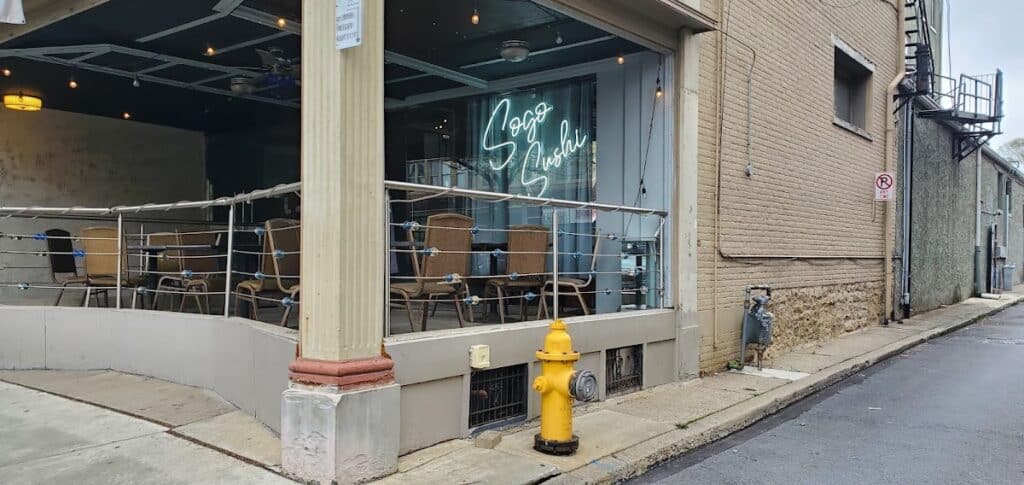 Exterior view of Sogo Sushi restaurant with outdoor seating and neon sign, located on a city street corner with brick building and fire hydrant nearby.