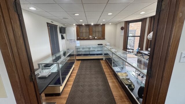 Interior view of a coin shop with glass display cases on both sides, wooden accents, and natural light from large windows, showcasing a clean and organized space.
