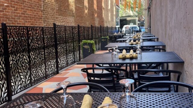 Outdoor dining area with black metal tables and chairs, set with napkins and glassware, in a narrow alleyway with brick and painted walls, ready for service.