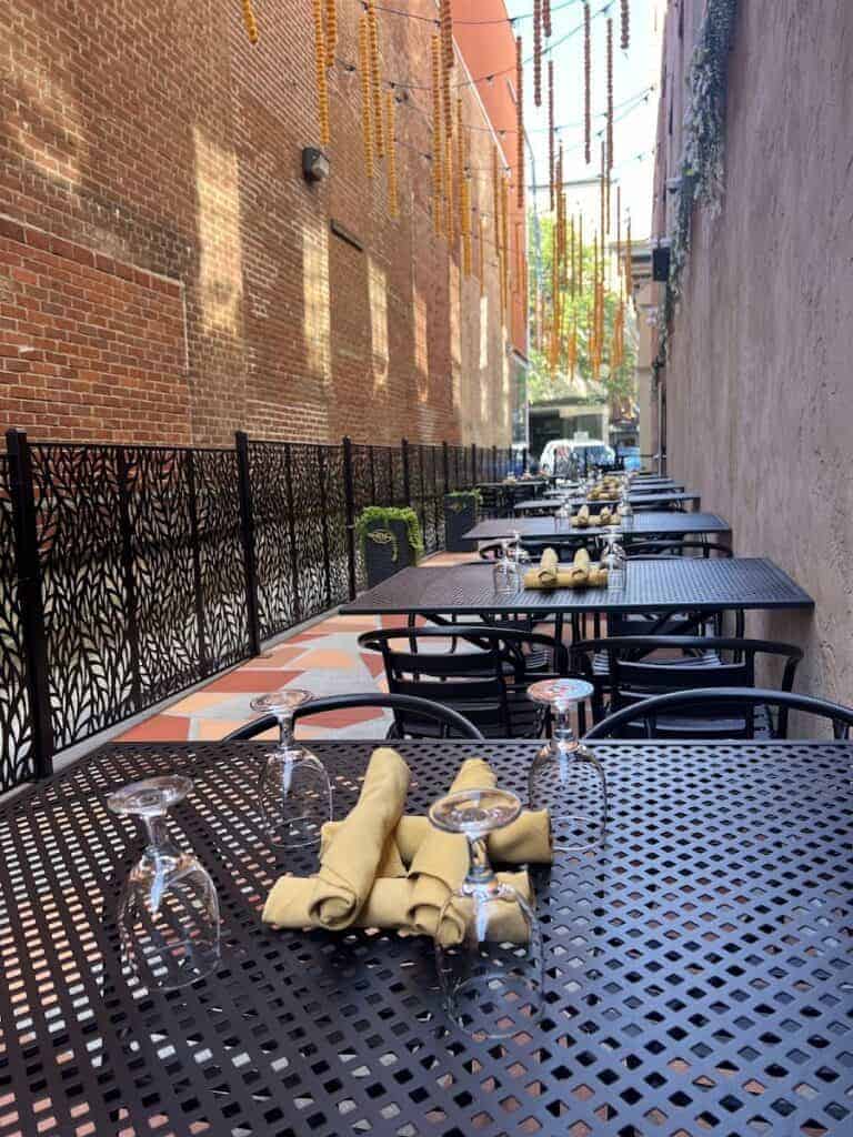 Outdoor dining area with black metal tables and chairs, set with napkins and glassware, in a narrow alleyway with brick and painted walls, ready for service.