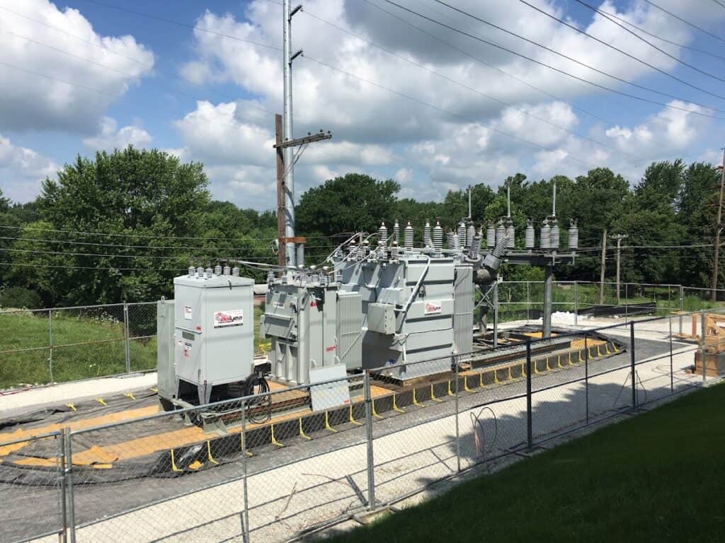 Electrical transformer station with power lines and green trees in the background under a partly cloudy sky.