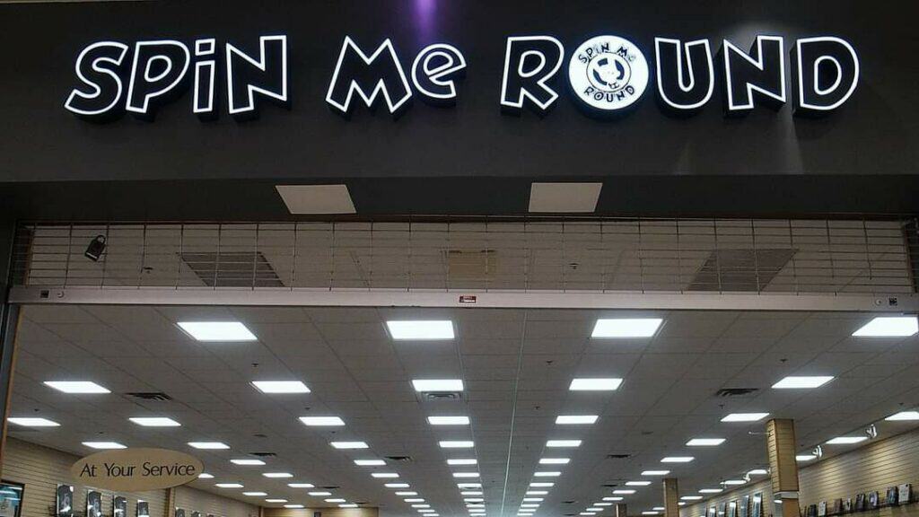 Sign for 'Spin Me Round' with bold black and white lettering above a brightly lit indoor space.