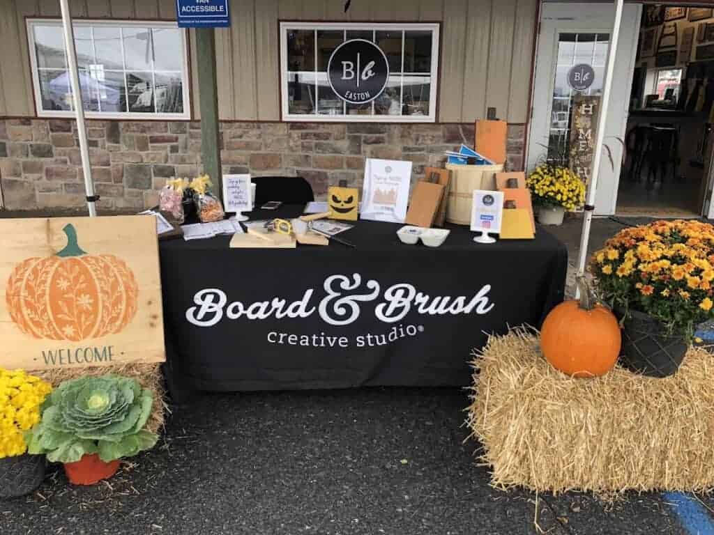 Outdoor display table at Board & Brush Creative Studio in Easton with pumpkins, flowers, and craft supplies for DIY woodworking projects.
