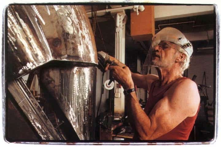 An elderly man with a white beard, wearing safety glasses and a sleeveless shirt, working on a large metallic sculpture in an art studio.