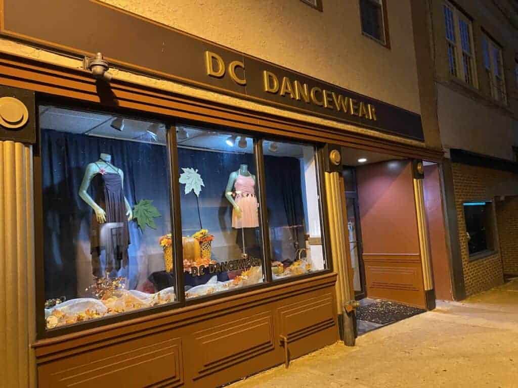 Exterior view of DC Dancewear store with window display of dance costumes and decorations, located on a city street at night.
