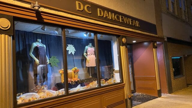 Exterior view of DC Dancewear store with window display of dance costumes and decorations, located on a city street at night.