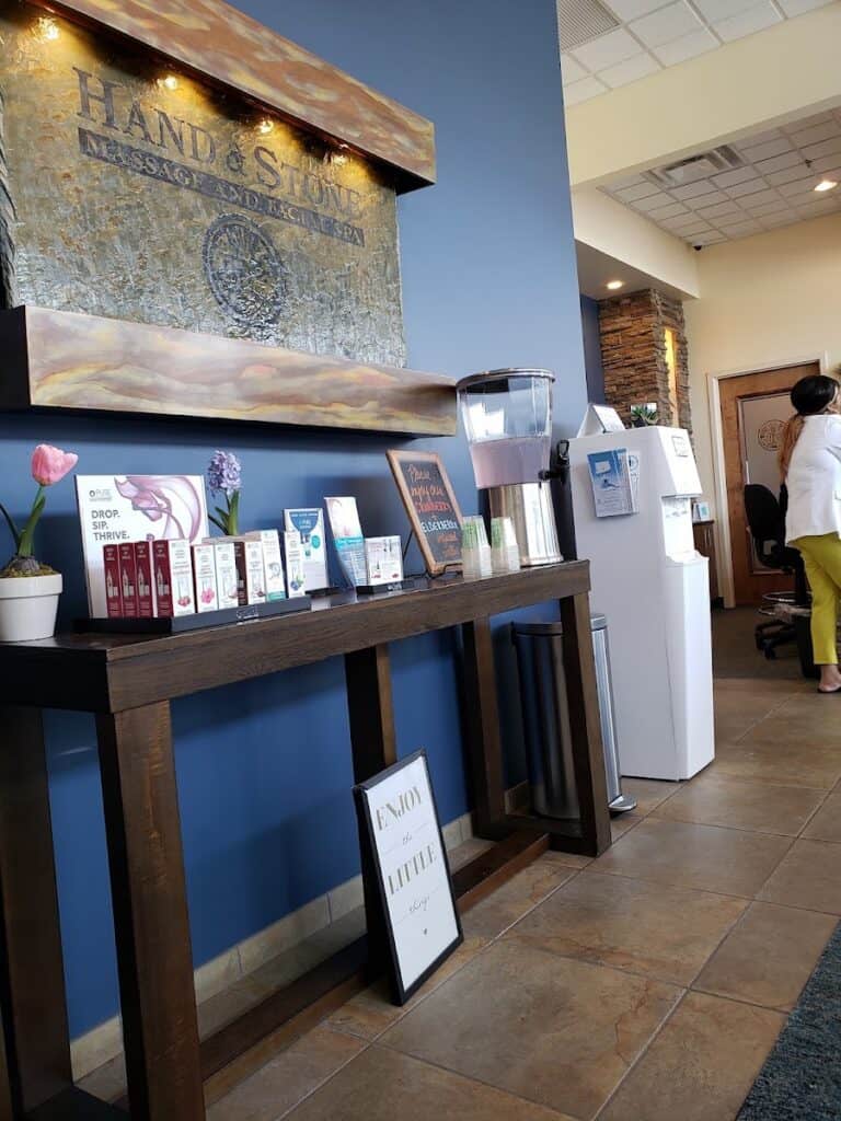 Interior view of Hand & Stone Massage and Facial Spa reception area with water dispenser, promotional materials, and a staff member in the background.