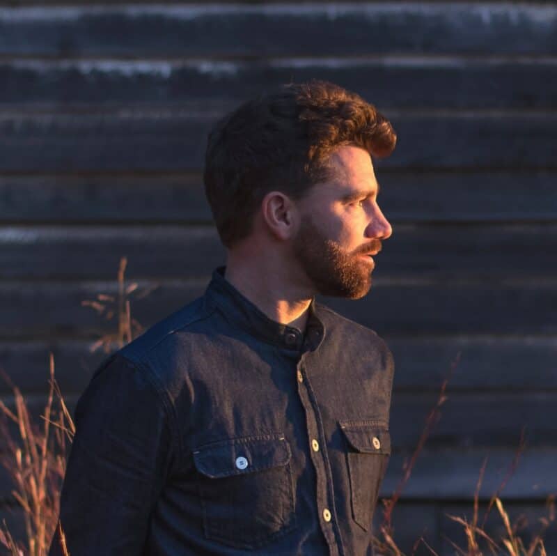 Portrait of a man with a beard and short hair, wearing a dark denim shirt, standing outdoors during sunset with a wooden background.