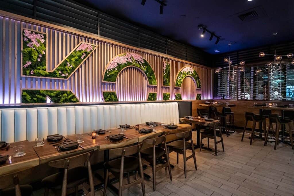 Modern restaurant interior with a large illuminated 'VICO' sign made of greenery and flowers, wooden accents, and a cozy dining area with tables and chairs.