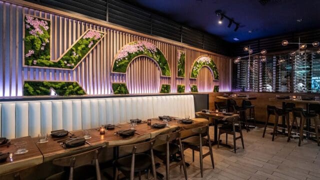 Modern restaurant interior with a large illuminated 'VICO' sign made of greenery and flowers, wooden accents, and a cozy dining area with tables and chairs.