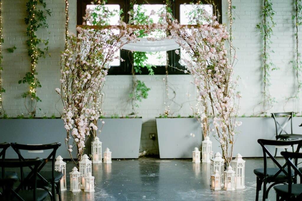 Elegant wedding ceremony setup with floral arch, lanterns, and chairs in a decorated indoor venue for Kraft & Co. Floral and Gifts.