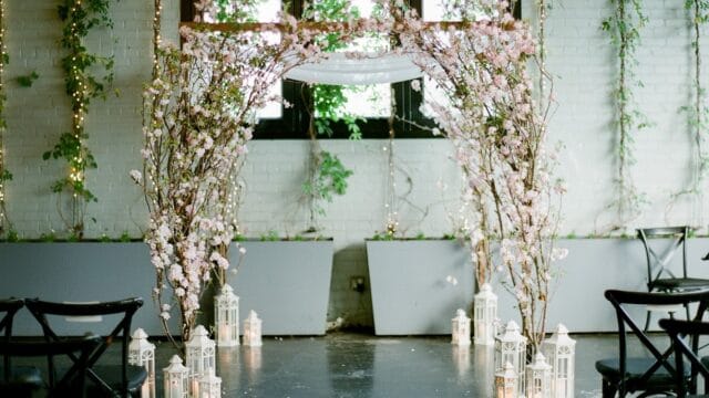 Elegant wedding ceremony setup with floral arch, lanterns, and chairs in a decorated indoor venue for Kraft & Co. Floral and Gifts.