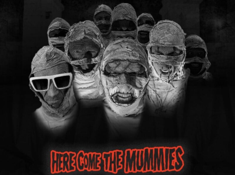 Promotional image for HERE COME THE MUMMIES at Musikfest Café featuring band members in mummy costumes with sunglasses and face paint, dark background, and red event title text.